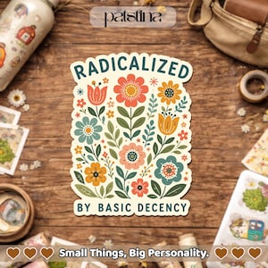 May include: A sticker with a floral design and the words "RADICALIZED BY BASIC DECENCY." The sticker features colorful flowers in shades of orange, blue, and pink, with green leaves. The sticker is on a wooden surface.