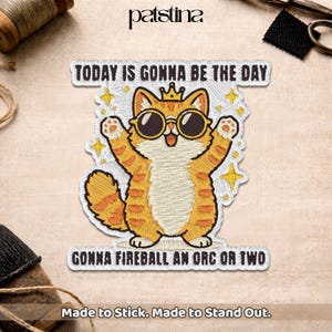 Peut inclure: Écusson brodé avec un chat de dessin animé portant des lunettes de soleil et une couronne. Le texte indique "TODAY IS GONNA BE THE DAY" et "GONNA FIREBALL AN ORC OR TWO". L'écusson est orange, blanc et jaune, avec les mots "Made to Stick. Made to Stand Out."
