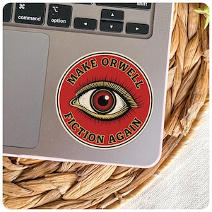 May include: A round sticker with the text "MAKE ORWELL FICTION AGAIN" in a circular design. The sticker features a red background with a detailed eye illustration in the center. The sticker is on a laptop.