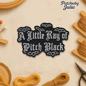 May include: A black patch with white embroidered text that reads "A Little Ray of Pitch Black." The patch is surrounded by bat silhouettes. The image also shows sewing supplies, including thread, scissors, and pins.