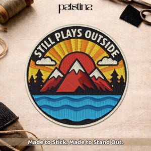 May include: Embroidered patch with a mountain and sunset design. The patch features the text "STILL PLAYS OUTSIDE" in white, with a red mountain, blue water, and yellow and orange sun rays. The patch is round and has the text "Made to Stick. Made to Stand Out."