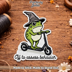 May include: Embroidered patch featuring a green frog wearing a witch hat and riding a black scooter. The patch has the text "Off to assess behavior" and "Made to Stick. Made to Stand Out."
