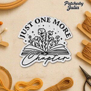 May include: A white and black embroidered patch with the text "JUST ONE MORE CHAPTER" surrounding an open book with floral elements. The patch is surrounded by sewing supplies, including thread and scissors.