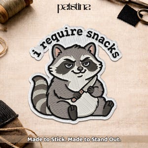 May include: Embroidered patch featuring a cartoon raccoon holding a knife, with the text "I require snacks" above. The patch is gray, white, and black, with the words "Made to Stick. Made to Stand Out." below.