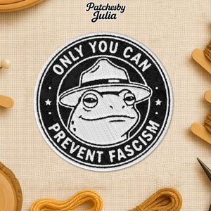 May include: Embroidered patch with a black border and a white frog wearing a ranger hat. The text "ONLY YOU CAN PREVENT FASCISM" is in a circular design around the frog. The patch is on a beige fabric background.