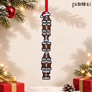 May include: A vertical hanging Christmas ornament featuring the text ANYNAME in brown football-textured letters with white stitching details. The ornament is topped with a red and white Santa hat and hangs from a red ribbon.