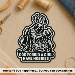 May include: Black and white embroidered patch featuring a hooded figure holding a skull and surrounded by ghosts. The text "GOD FORBID A GIRL HAVE HOBBIES" is displayed below the figure. The patch is likely for clothing or accessories.