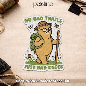 May include: Embroidered patch featuring a cartoon bear with glasses, a hat, and a backpack, holding a walking stick. The patch reads "NO BAD TRAILS JUST BAD KNEES" with butterflies and flowers. The text "Made to Stick. Made to Stand Out." is also included.