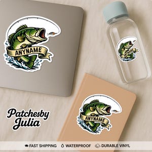 May include: A sticker featuring a green and yellow bass fish with an open mouth, a fishing rod, and the text "ANYNAME" on a banner. The sticker is displayed on a laptop, a water bottle, and a notebook.
