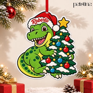 May include: A hanging Christmas ornament featuring an embroidered green dinosaur wearing a red Santa hat with the name Roberto written in red script. The dinosaur holds a decorated Christmas tree with red and blue ornaments and a yellow star.
