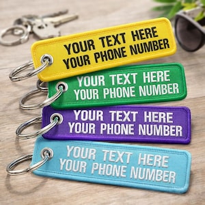 May include: Four rectangular keychains in yellow, green, purple, and light blue, each with silver key rings. The text "YOUR TEXT HERE YOUR PHONE NUMBER" is embroidered in white on each keychain. The keychains are on a wooden surface.