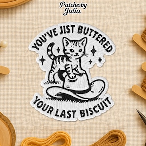 May include: Embroidered patch featuring a cartoon cat standing on a hat, with the text "You've Jist Buttered Your Last Biscuit". The patch is white with black details and a cream-colored background.