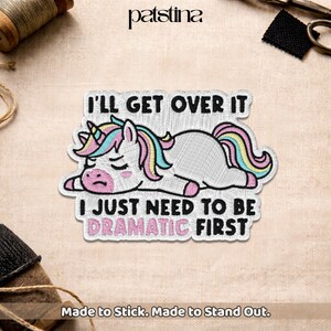 May include: Embroidered patch featuring a white unicorn with a colorful mane and tail, lying down. The patch has the text "I'LL GET OVER IT I JUST NEED TO BE DRAMATIC FIRST". The patch is made to stick and stand out.