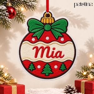 May include: A red and white embroidered Christmas ornament shaped like a bauble, featuring a green bow at the top, a central cream-coloured band with the name Mia in red script, and three green pine trees at the bottom.