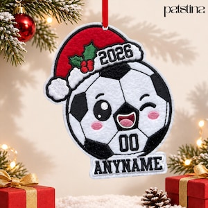 May include: A white and black soccer ball ornament featuring a red Santa hat with green holly and red berries. The ball has a winking face, the year 2026, and a white banner at the bottom with the text ANYNAME in black embroidery.