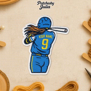 May include: Embroidered patch featuring a baseball player in a blue uniform with yellow accents, swinging a bat. The patch includes the number 9 and the text "YOUR NAME". The patch is on a beige background.
