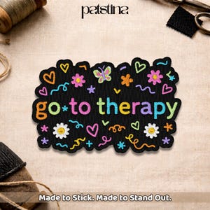 May include: Black embroidered patch with the words "go to therapy" in colorful letters. The patch is decorated with embroidered hearts, flowers, butterflies, and stars. The text "Made to Stick. Made to Stand Out." is at the bottom.