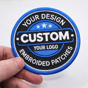 Peut inclure: Écusson brodé rond avec un design bleu et noir. Le patch porte les mots "YOUR DESIGN", "CUSTOM", "YOUR LOGO" et "EMBROIDERED PATCHES" en lettres blanches. Il a une bordure bleue et trois petites étoiles.