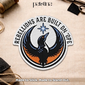 May include: Embroidered patch with a circular design featuring a stylized bird with outstretched wings. The patch has a black, blue, and orange color scheme, with the text "REBELLIONS ARE BUILT ON 'OPE" around the edge. The text "Made to Stick. Made to Stand Out." is at the bottom.