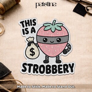 Peut inclure: Un écusson brodé représentant une fraise rose avec un masque gris, tenant un sac d'argent beige avec un signe dollar. Le texte THIS IS A STROBBERY est inscrit en lettres noires. Le motif inclut une tige verte et des membres noirs.