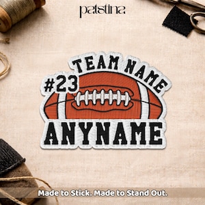 May include: An embroidered patch featuring a brown football with white laces, surrounded by black text that reads TEAM NAME, #23, and ANYNAME. The design is outlined in white thread and has a textured appearance suitable for application to fabric items.