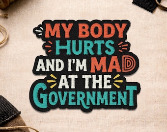 Puede incluir: Parche bordado negro con el texto "MY BODY HURTS AND I'M MAD AT THE GOVERNMENT" en naranja, turquesa y blanco. El parche tiene un borde negro y las palabras "Made to Stick. Made to Stand Out."
