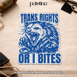May include: Embroidered patch with the text "TRANS RIGHTS OR I BITES" in blue and white. The patch features a detailed illustration of an opossum with an open mouth, showcasing its teeth. The patch is made to stick.