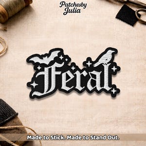 May include: Black and white embroidered patch with the word "Feral" in gothic lettering, featuring a bat and a bird. The patch is surrounded by stars. Text at the bottom reads "Made to Stick. Made to Stand Out."