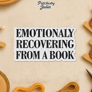 May include: Embroidered patch with the text "EMOTIONALY RECOVERING FROM A BOOK" in black on a white background. The patch has a decorative border and is surrounded by craft supplies, including thread and scissors.