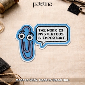 May include: Embroidered patch featuring a blue paperclip with googly eyes and a speech bubble. The speech bubble contains the text "THE WORK IS MYSTERIOUS & IMPORTANT." The patch is on a beige fabric background. The text "Made to Stick. Made to Stand Out." is at the bottom.