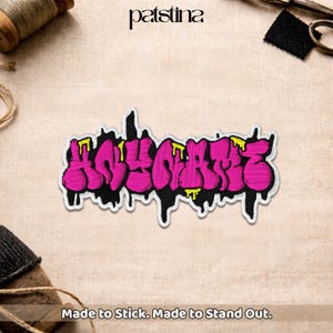 May include: Embroidered patch with the word "HOLLABACK" in a graffiti style. The patch features bright pink letters outlined in black with yellow accents, and a white border. The text "Made to Stick. Made to Stand Out." is at the bottom.