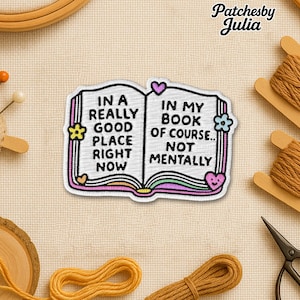 May include: Embroidered patch depicting an open book with the text "IN A REALLY GOOD PLACE RIGHT NOW" and "IN MY BOOK OF COURSE... NOT MENTALLY". The patch has a white border and colorful accents. The patch is surrounded by sewing supplies.