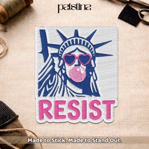 May include: Embroidered patch featuring the Statue of Liberty wearing heart-shaped sunglasses and blowing a bubblegum bubble. The word "RESIST" is embroidered in pink below the image. The patch is white with blue and pink accents. Text at the bottom reads "Made to Stick. Made to Stand Out."