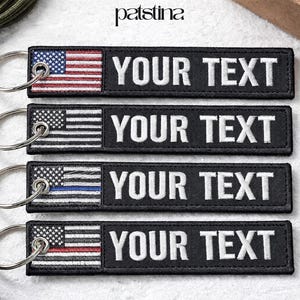 May include: Black rectangular keychains with the American flag design and the text "YOUR TEXT" in white. The keychains feature different flag variations, including a standard color flag, a black and white flag, a thin blue line flag, and a thin red line flag.