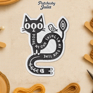 May include: Embroidered black and white patch featuring a stylized cat with the text "Doing my own thing in my own time." A small bird is perched on the cat's curved tail. The patch is surrounded by sewing supplies.