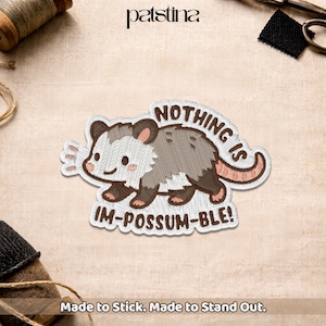 May include: Embroidered patch featuring a cartoon opossum with the text "NOTHING IS IM-POSSUM-BLE!". The patch has a white, gray, and brown color scheme with a pink tail and ears. The patch is on a beige fabric.