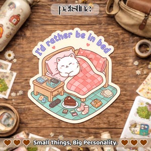 May include: A die-cut sticker featuring a cartoon cat in bed, with the text "I'd rather be in bed." The sticker includes a nightstand with a game console, remote, and a slice of cake. The sticker also has the text "Small Things, Big Personality."