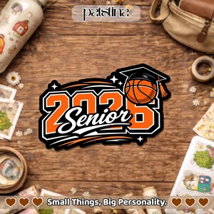 Pu&ograve; includere: Un adesivo arancione, nero e bianco con la scritta "2023 Senior" e un pallone da basket con un cappello di laurea. L'adesivo ha un contorno nero ed &egrave; su una superficie di legno. Il testo "Small Things, Big Personality" &egrave; in basso.