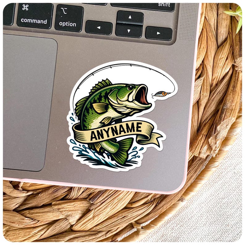 May include: A sticker featuring a green and black bass fish jumping out of the water with a fishing lure. The sticker has a banner that says "ANYNAME" in black text. The sticker is on a laptop.