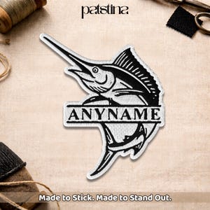 May include: Embroidered patch with a black and white marlin fish design and the text "ANYNAME" in a white banner. The patch is on a beige fabric background. The text "Made to Stick. Made to Stand Out." is at the bottom.
