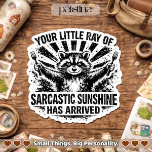 Puede incluir: Una pegatina en blanco y negro con un mapache sosteniendo tenedores y el texto "YOUR LITTLE RAY OF SARCASTIC SUNSHINE HAS ARRIVED." La pegatina también incluye el texto "Small Things, Big Personality."