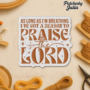 May include: Embroidered patch with the text "AS LONG AS I'M BREATHING I'VE GOT A REASON TO PRAISE the LORD" in brown thread on a white background. The patch is surrounded by craft supplies, including thread spools, scissors, and a wooden embroidery hoop.