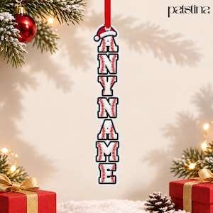 May include: A vertical hanging Christmas ornament featuring the text ANY NAME in block letters styled like baseballs with red stitching and black outlines. The top letter A is topped with a small red and white Santa hat. The ornament hangs from a red ribbon.