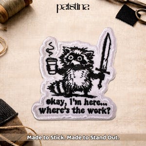 May include: Embroidered patch featuring a raccoon holding a coffee cup and a sword. The raccoon is black and white with a white border. Text on the patch reads "okay, I'm here... where's the work?" The patch is designed to stick.