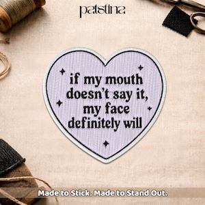 May include: A heart-shaped embroidered patch in light lavender with a white border. The patch features black text that reads, "if my mouth doesn't say it, my face definitely will," accented by four small black four-pointed star symbols.