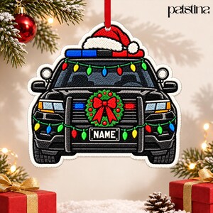 May include: A flat, die-cut Christmas ornament shaped like a black police cruiser. The vehicle features a red Santa hat on the roof, a green wreath with a red bow on the grille, and colorful string lights. A license plate reads NAME.