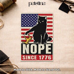 May include: Embroidered patch featuring a black cat with a crown, set against an American flag design. The word "NOPE" is displayed above "SINCE 1776" in a bold, red and white font. The patch is rectangular and has the text "Made to Stick. Made to Stand Out."