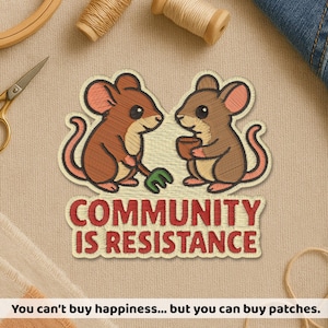 May include: Embroidered patch featuring two cartoon mice facing each other. The patch reads "COMMUNITY IS RESISTANCE" in red thread. One mouse holds a green object, the other a brown cup. The patch is on a beige fabric.