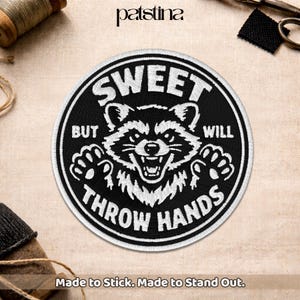 May include: A round, black and white embroidered patch featuring a raccoon with the text "SWEET BUT WILL THROW HANDS." The patch also includes the text "Made to Stick. Made to Stand Out."