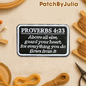 May include: Black rectangular patch with white embroidered text from Proverbs 4:23: "Above all else, guard your heart, for everything you do flows from it." The patch has a white border and is surrounded by sewing supplies.